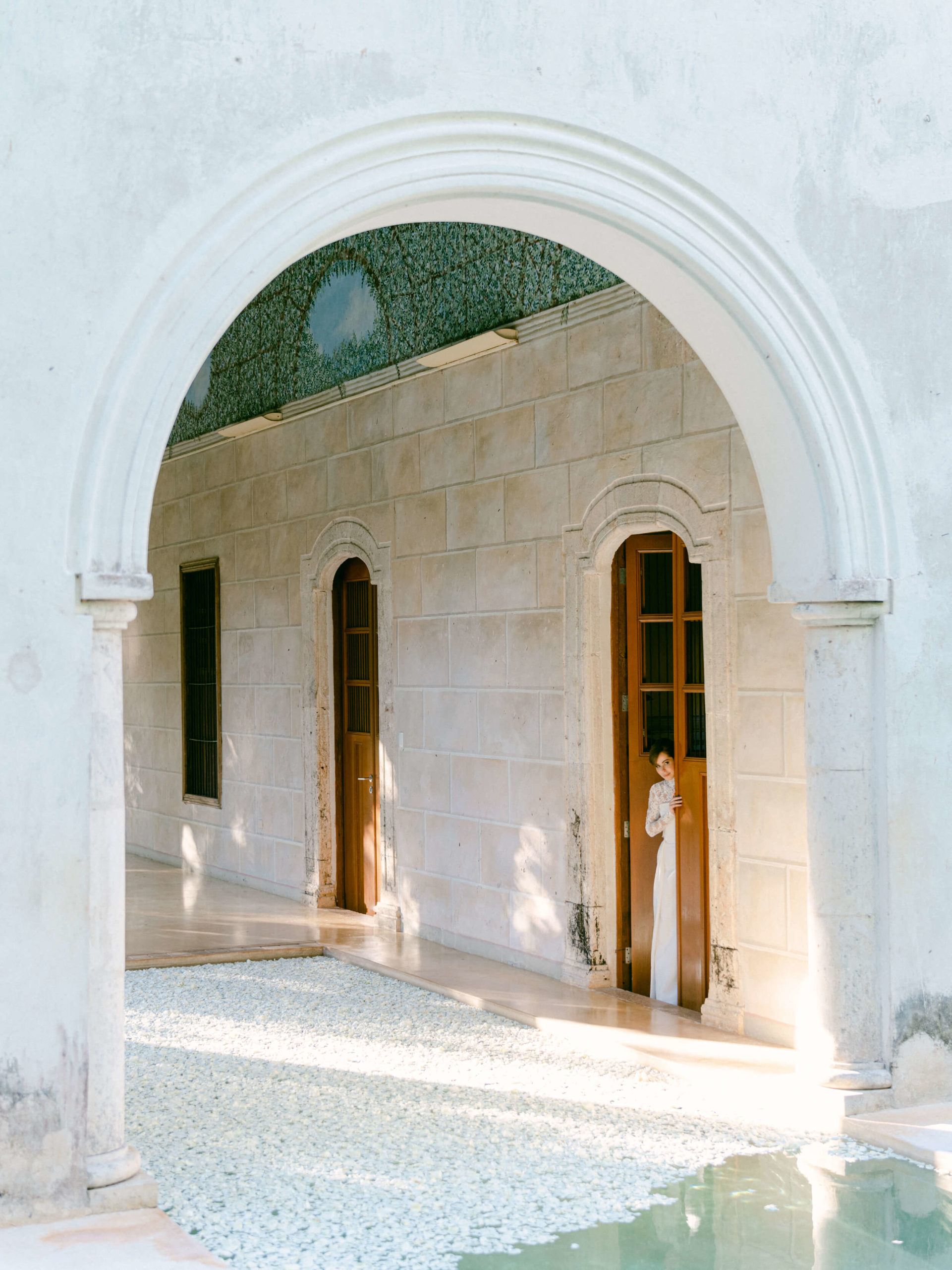 Bride peeking out from behind a door of a historical villa in her wedding dress