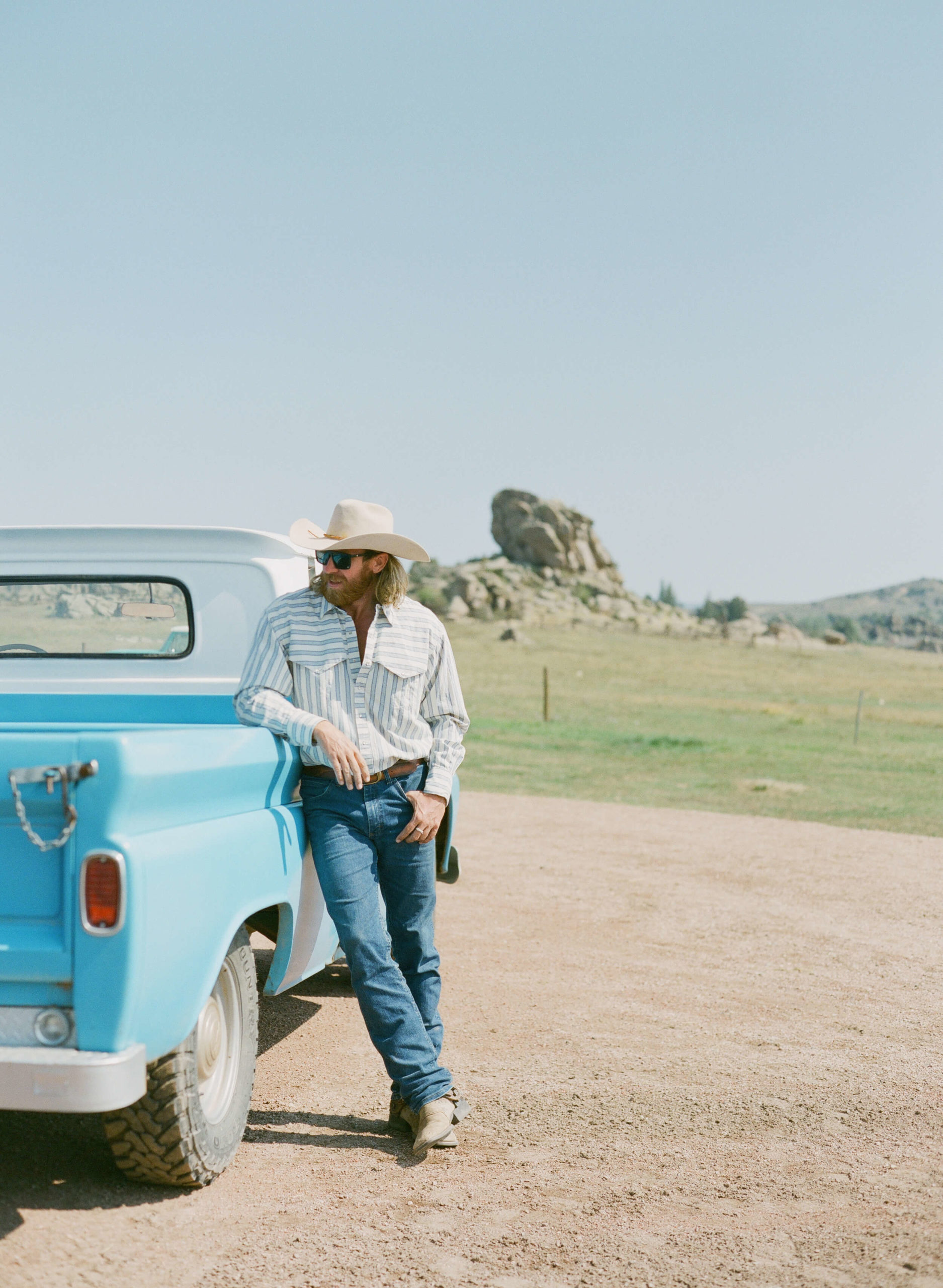 Man in western garb leaning against a vintage blue pickup truck