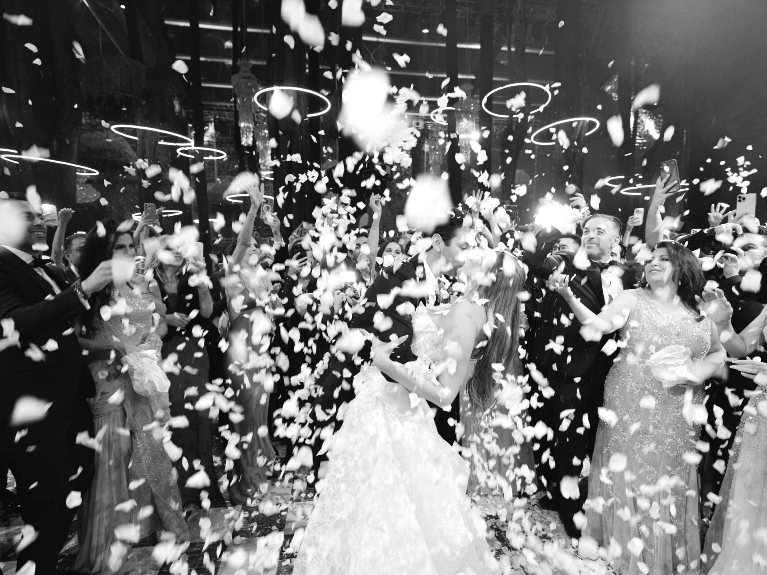 Black and white shot of Ashley and Brian kissing as guests cheer and flower petals fall from above